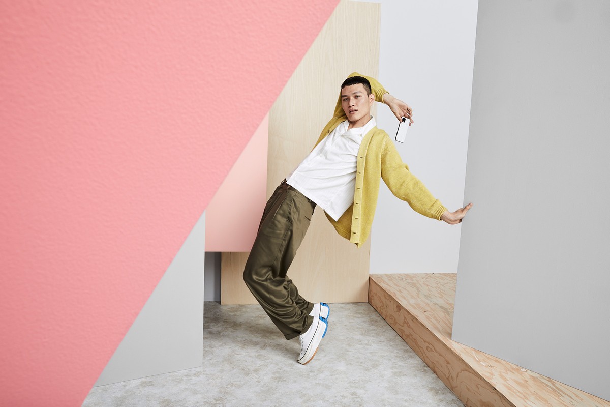 A person in olive pants and yellow cardigan strikes a pose between angled walls in front of a concrete floor in a studio setting.