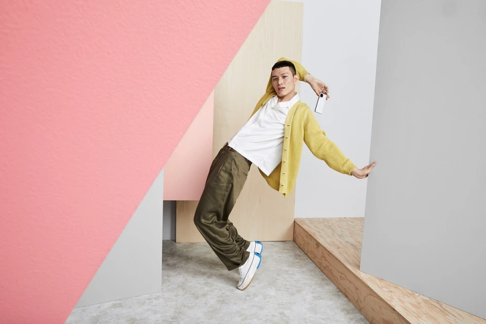 A person in olive pants and yellow cardigan strikes a pose between angled walls in front of a concrete floor in a studio setting.