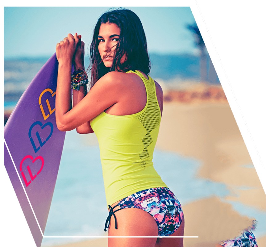 Woman in activewear holds a surfboard on a beach. She wears a yellow top and patterned bikini bottom, with a blue sky background.