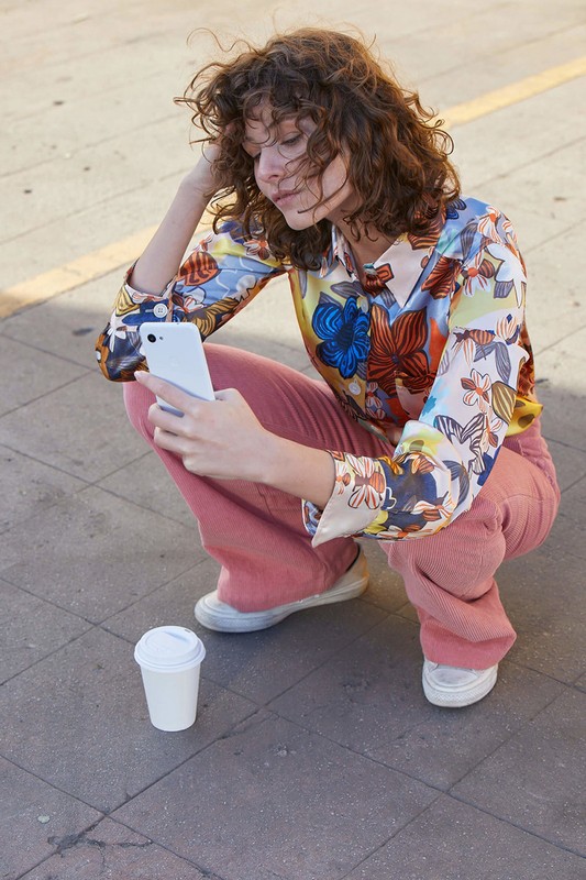 Woman with curly brown hair sitting in urban setting, looking at smartphone. Dressed in floral top, pink pants, and white sneakers.