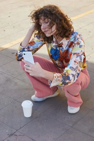 Woman with curly brown hair sitting in urban setting, looking at smartphone. Dressed in floral top, pink pants, and white sneakers.
