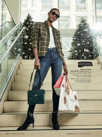 A fashionable Black woman stands on stairs with Christmas trees in a department store, wearing a blazer, jeans, and sunglasses.