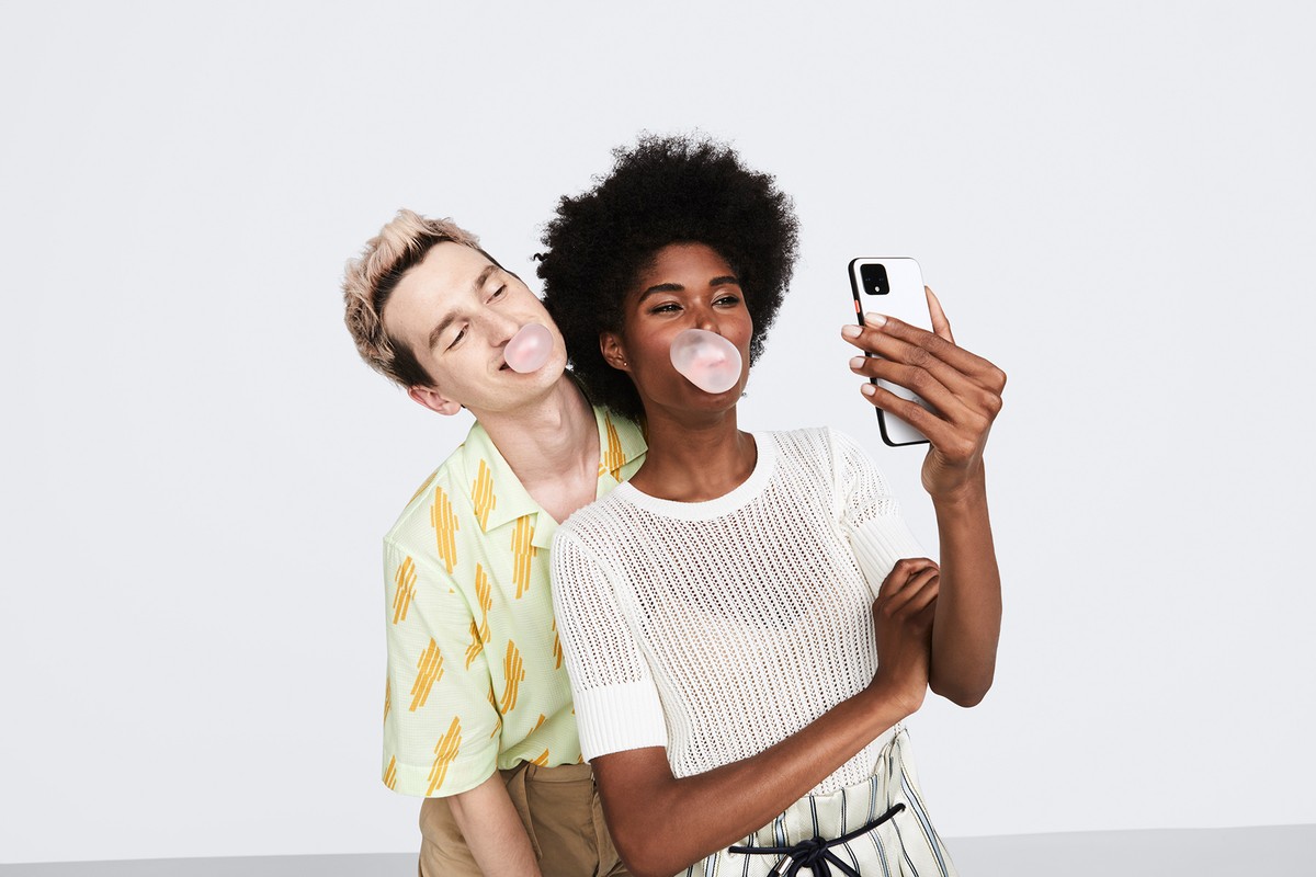 Two people, a man and a woman, pose indoors blowing bubble gum bubbles while the woman holds up a smartphone to take a selfie.