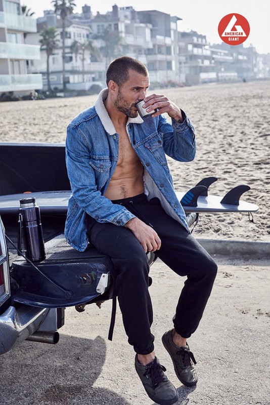 Man in denim jacket drinks coffee on a beach near a truck with a surfboard. Buildings and sky are in the background.