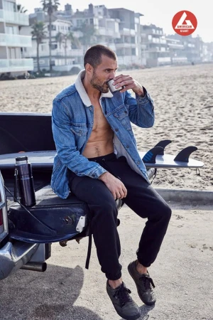 Man in denim jacket drinks coffee on a beach near a truck with a surfboard. Buildings and sky are in the background.  