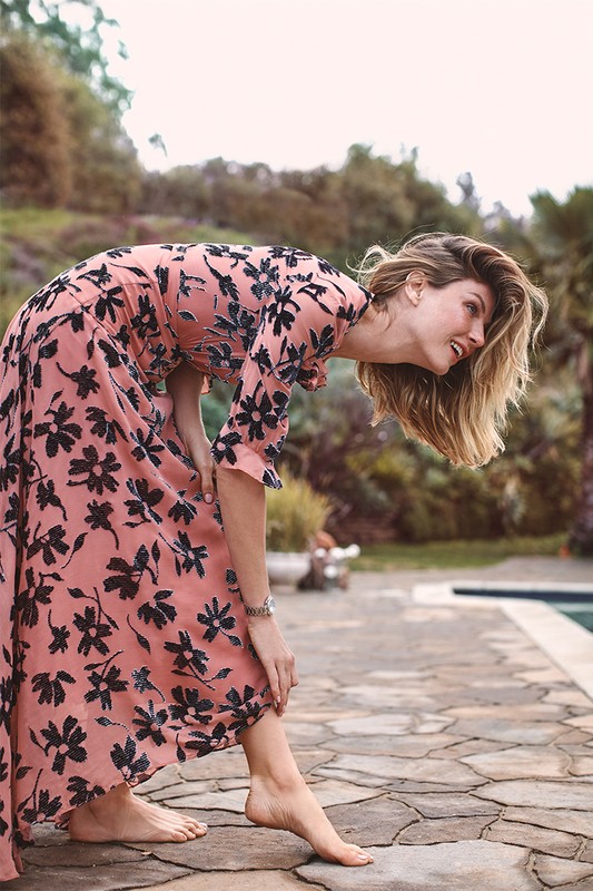A woman in a long floral dress poses barefoot by a pool, leaning forward with her back arched and head down, set against a lush backdrop.