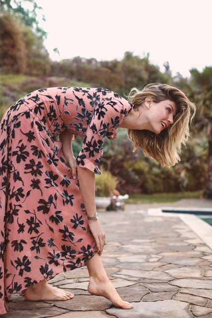 A woman in a long floral dress poses barefoot by a pool, leaning forward with her back arched and head down, set against a lush backdrop.