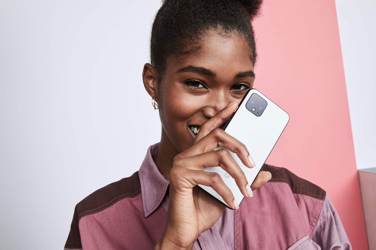 Smiling woman holds white smartphone indoors against a colorful background, showcasing modern technology and casual fashion.