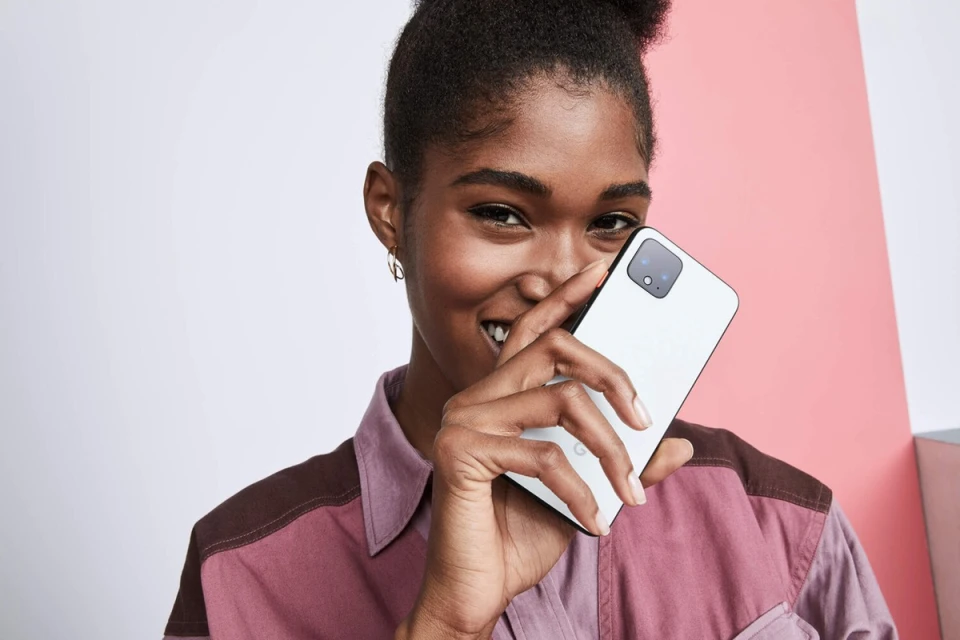 Smiling woman holds white smartphone indoors against a colorful background, showcasing modern technology and casual fashion.