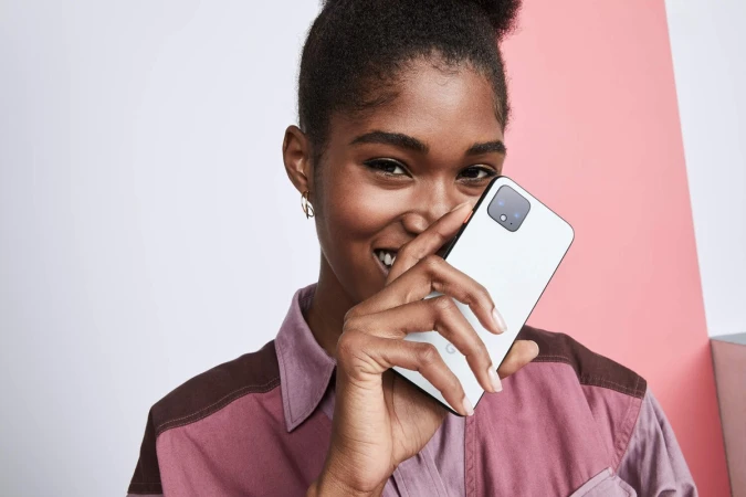 Smiling woman holds white smartphone indoors against a colorful background, showcasing modern technology and casual fashion.