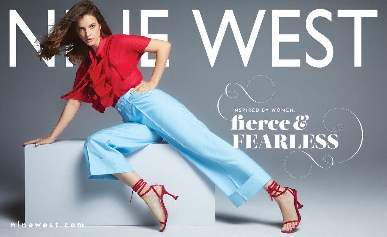 Barbara Palvin in a Nine West ad wearing a red blouse, light blue pants, and red heels, sitting on a white cube against a gray background.