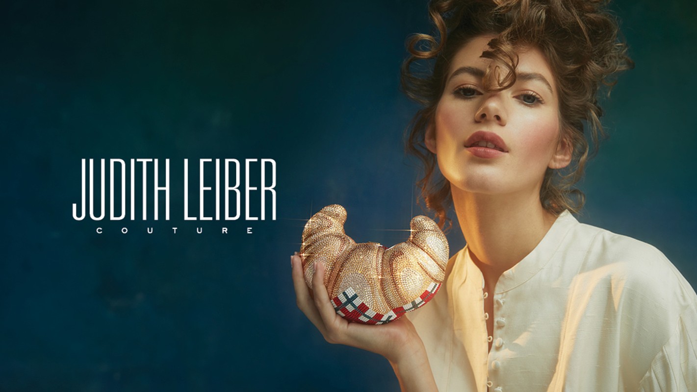 Woman holding a glittering Judith Leiber Couture croissant clutch against a teal background in a studio-lit beauty and fashion shot.