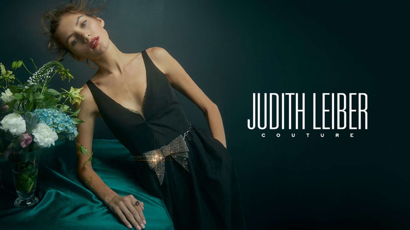 Couture fashion shot: a model in a black dress with a sparkling bow belt, leaning on a green velvet surface with a bouquet of flowers.