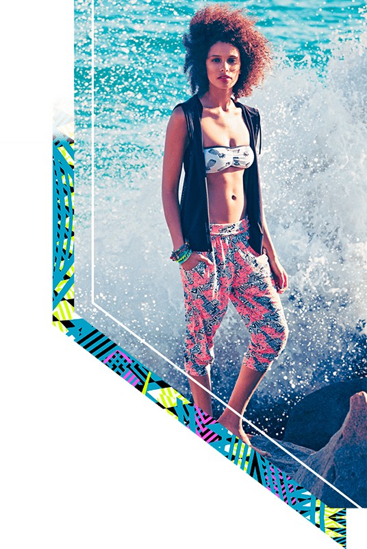 A young woman poses on rocks by the ocean, wearing a bikini top, black vest, and patterned cropped pants as a wave crashes behind her.