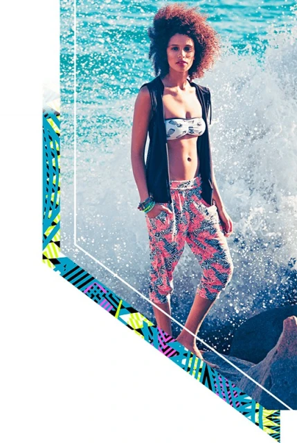 A young woman poses on rocks by the ocean, wearing a bikini top, black vest, and patterned cropped pants as a wave crashes behind her.