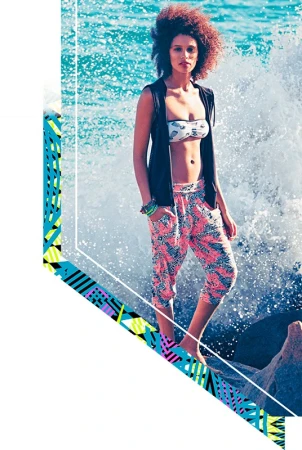 A young woman poses on rocks by the ocean, wearing a bikini top, black vest, and patterned cropped pants as a wave crashes behind her.