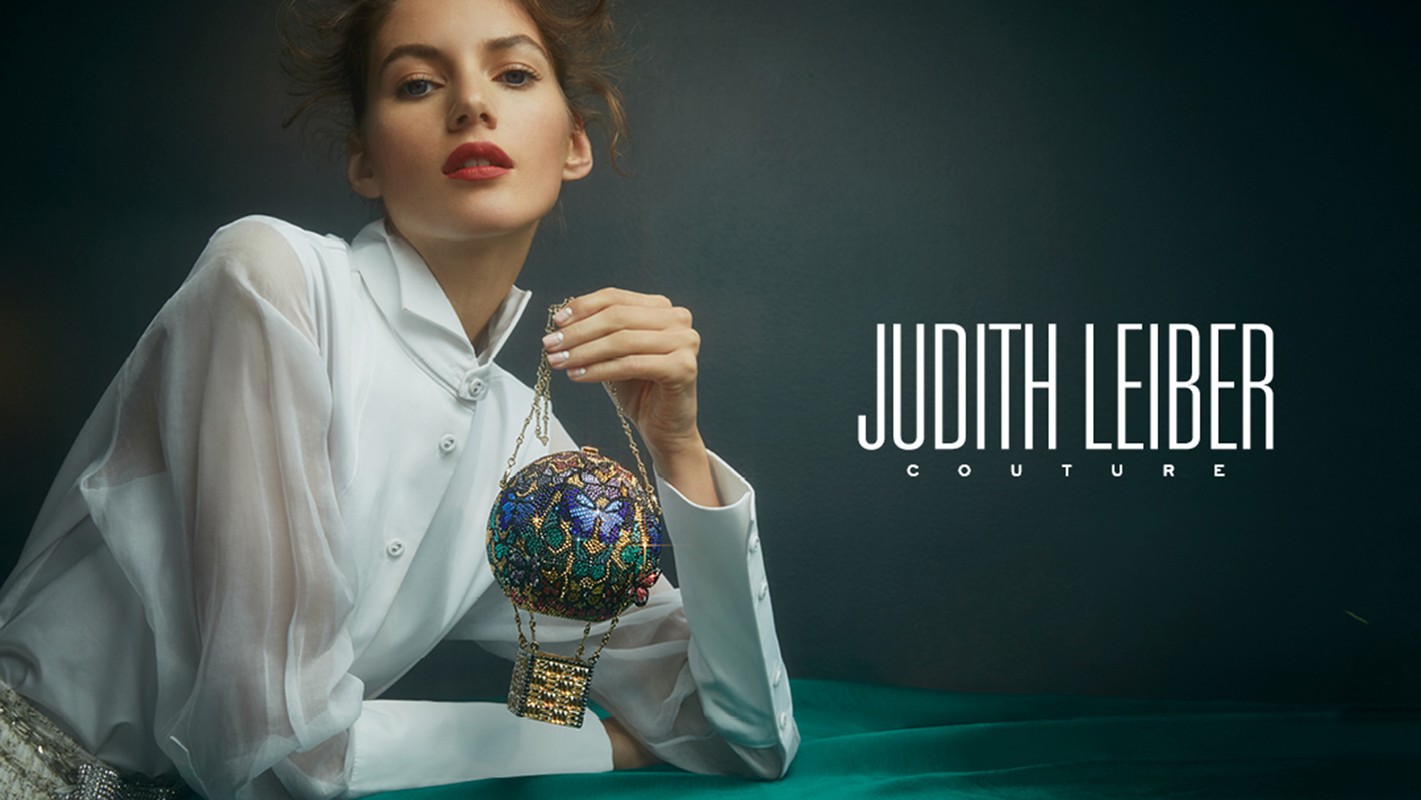 Fashion image of a poised female model holding a designer Judith Leiber Couture beaded handbag against a dark background.