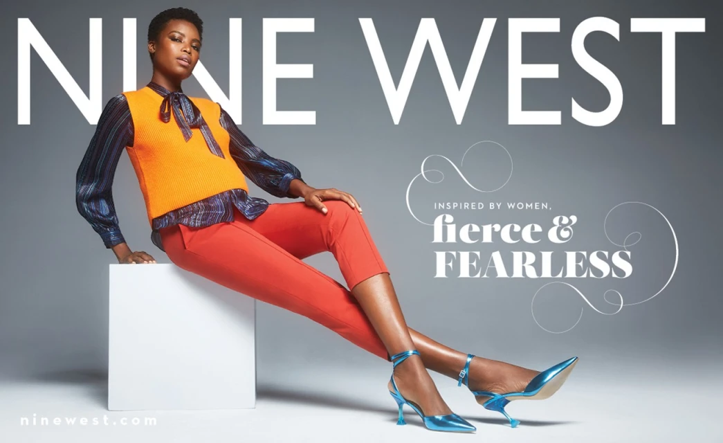 Nine West fashion campaign featuring a Black woman in an orange sweater vest, capri pants, and blue heels, sitting on a white block.