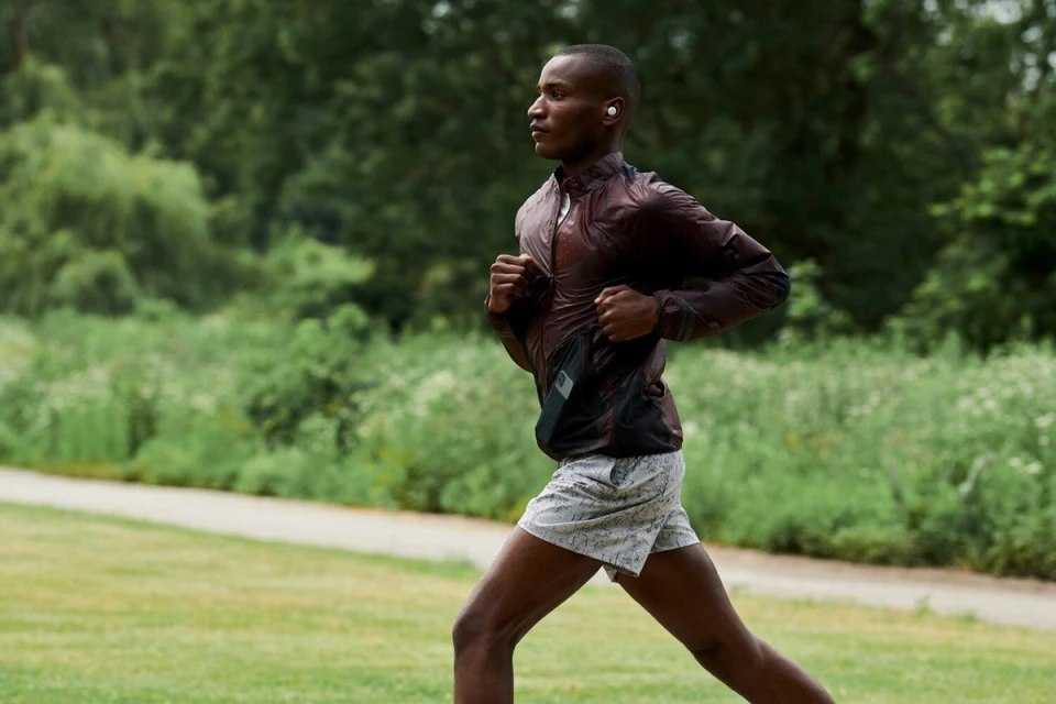 An African American man runs on a grassy path wearing a brown jacket and shorts, with trees and foliage in the background.