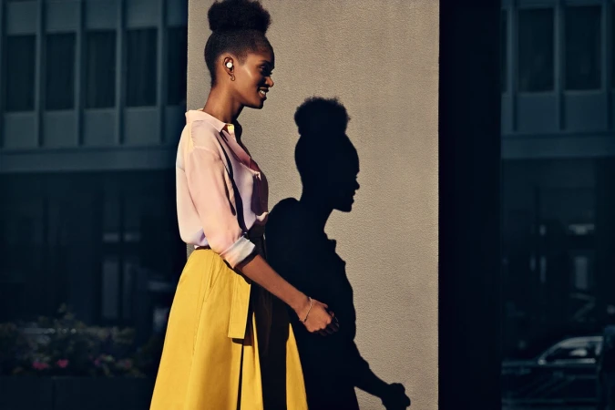 Black woman in a pink blouse and yellow skirt walking past a concrete pillar with her shadow cast on it.