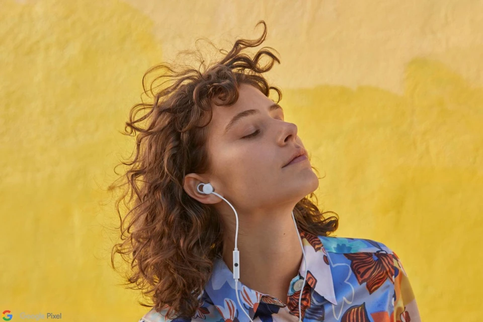 Woman with closed eyes listening to wired earbuds against a bright yellow background, wearing a patterned shirt with an upward gaze.