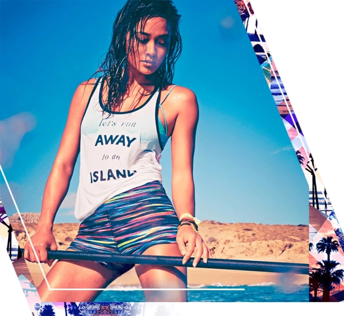 A young woman stands on a paddleboard in the ocean, wearing a white tank top and colorful shorts against a backdrop of blue sky.