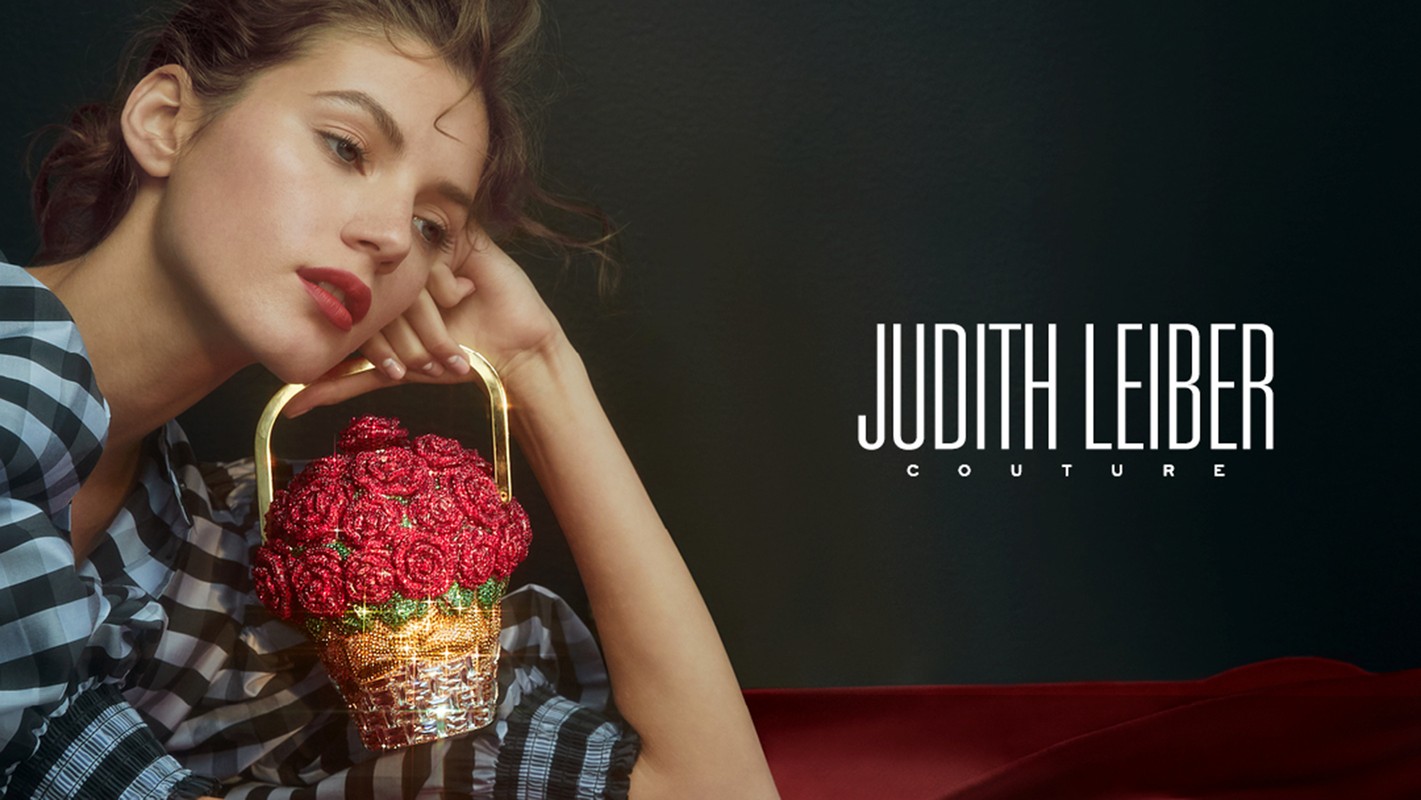 A woman models with a rose-themed Judith Leiber Couture purse, photographed in a studio against a dark background with red fabric.