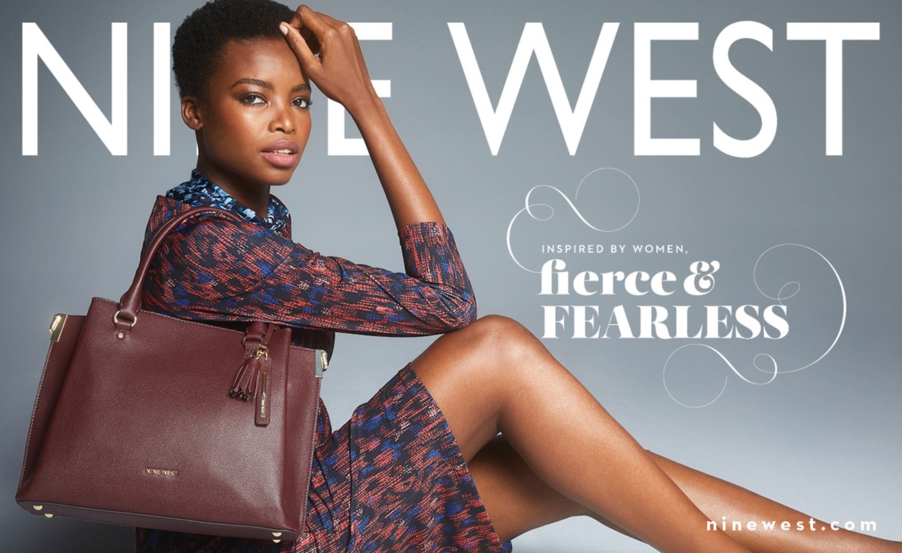 A dark-skinned female model poses with a burgundy Nine West handbag against a grey background, showcasing a sophisticated fashion aesthetic.