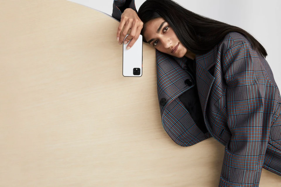 Brunette woman leans head on light-colored wood, wearing plaid jacket with a hand holding a white smartphone. Light-colored wall.