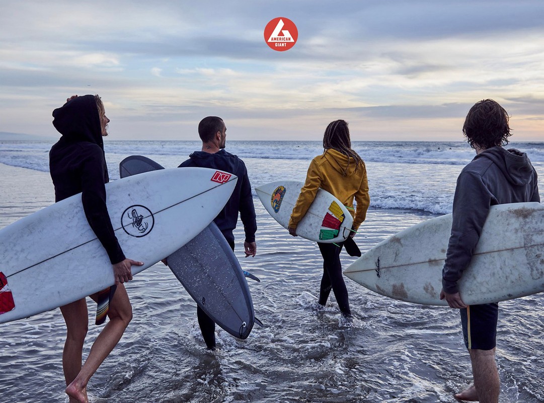 Group of four people walking into ocean with surfboards on a beach at sunset with an American Giant logo in the background.