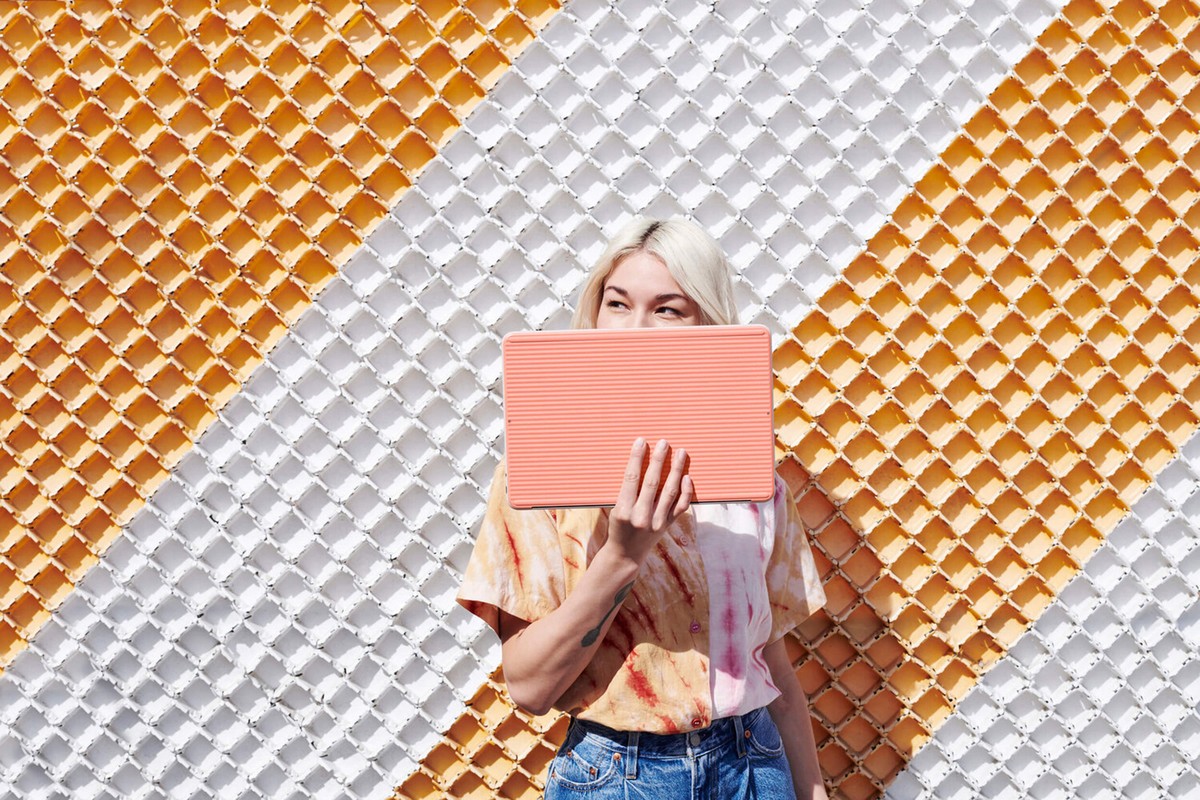 Woman with platinum blonde hair holds a striped peach-colored tablet in front of her face. She is wearing a tie-dye shirt and jeans.