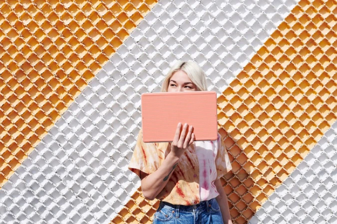 Woman with platinum blonde hair holds a striped peach-colored tablet in front of her face. She is wearing a tie-dye shirt and jeans.