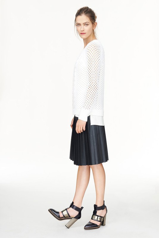 Full-length shot of a woman in a white sweater, black pleated skirt, and black high-heeled shoes against a plain white backdrop.