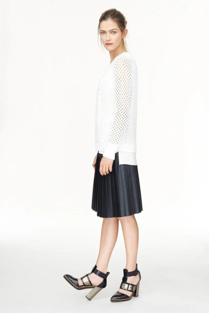 Full-length shot of a woman in a white sweater, black pleated skirt, and black high-heeled shoes against a plain white backdrop.