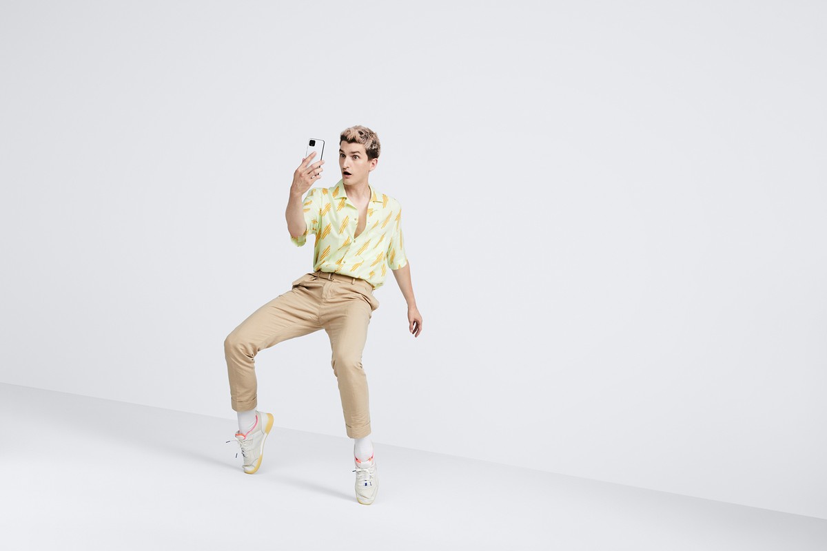 A young man with bleached blond hair poses in a studio setting, holding a smartphone and looking surprised in a light-hearted fashion shot.