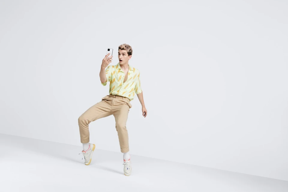 A young man with bleached blond hair poses in a studio setting, holding a smartphone and looking surprised in a light-hearted fashion shot.