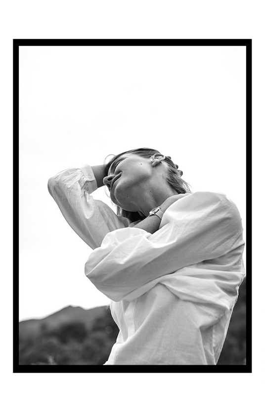 Monochrome portrait of a woman in a white top with her arms up, set against an outdoor background and framed in black and white.