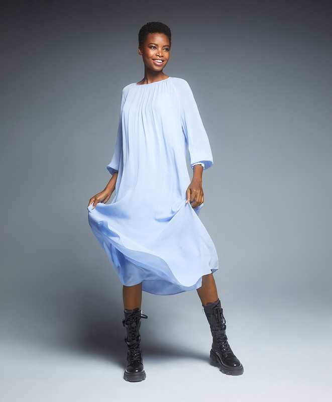 Full-length shot of an African-American woman wearing a light blue dress and black boots, posing in a studio with a grey background.