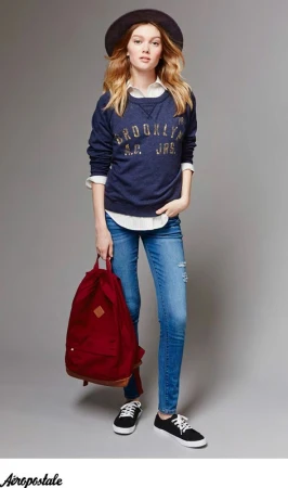 Model in dark blue sweatshirt and skinny jeans holding a red backpack against a grey seamless background