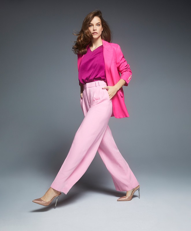 Elegant woman in pink suit walks confidently against gray backdrop. She wears a magenta top and clear heels in a studio fashion shot.
