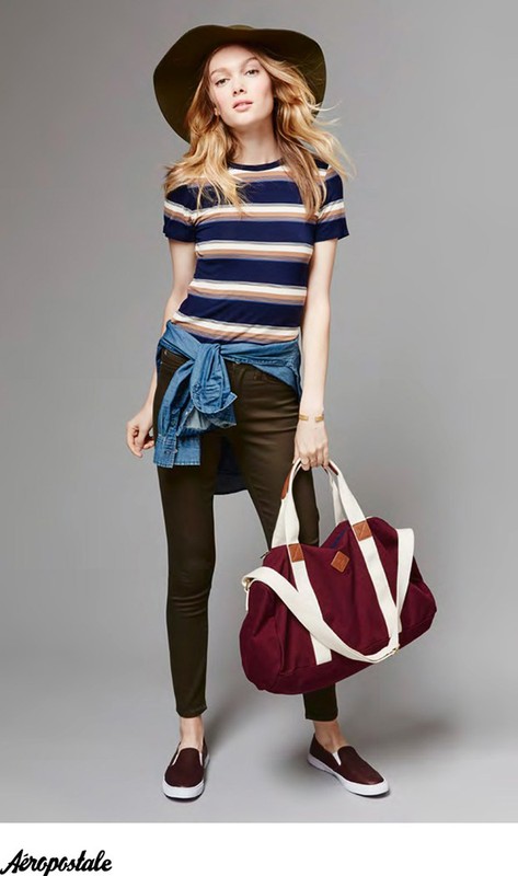 Model wearing striped t-shirt and wide-brim hat holding a large burgundy duffel bag against a grey seamless background