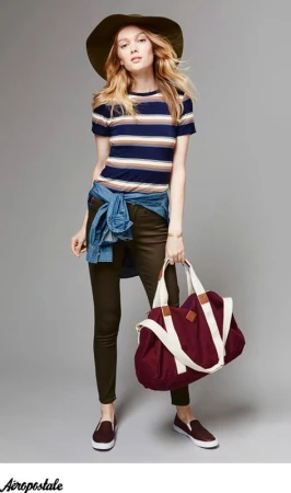 Model wearing striped t-shirt and wide-brim hat holding a large burgundy duffel bag against a grey seamless background