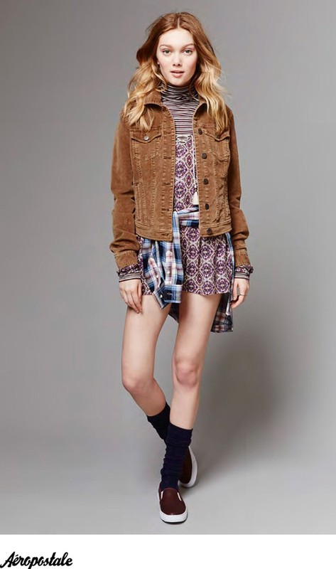 A young woman in a brown corduroy jacket, patterned dress, and leg warmers poses against a gray backdrop, showcasing a casual fashion style.