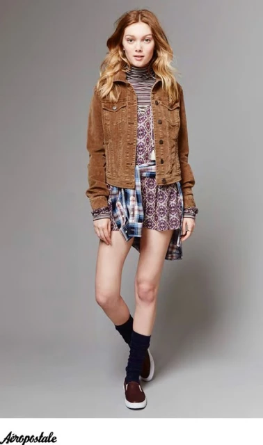 A young woman in a brown corduroy jacket, patterned dress, and leg warmers poses against a gray backdrop, showcasing a casual fashion style.