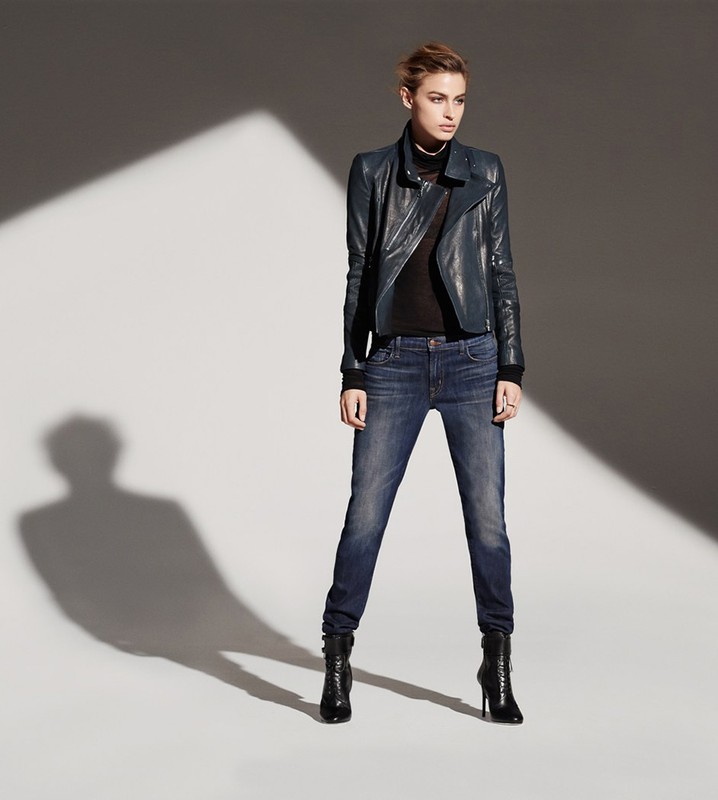 Model in dark blue leather jacket and denim jeans standing against a grey studio wall with dramatic diagonal light.