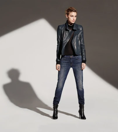 Model in dark blue leather jacket and denim jeans standing against a grey studio wall with dramatic diagonal light.