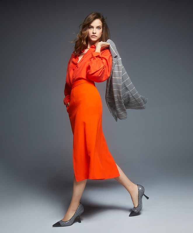 A woman in a red blouse and skirt strides confidently in a studio, carrying a gray plaid blazer over her shoulder, fashion-forward.