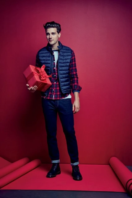 Young man in a puffer vest holds a red gift box, standing on a red surface against a matching background in a studio shot.