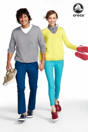 A fashionable couple wears Crocs shoes and apparel while walking hand-in-hand against a white background, showcasing casual style and comfort.