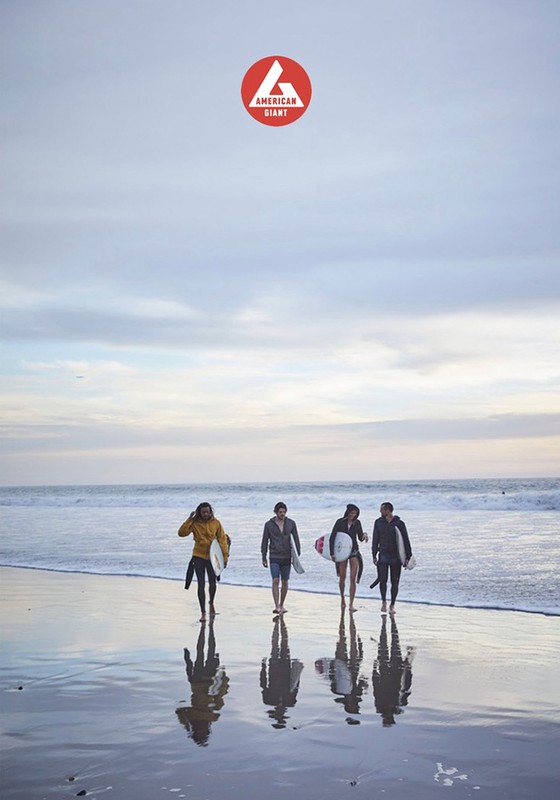 Four surfers walking on wet sand at beach toward the ocean with surfboards; American Giant logo at top center on a cloudy day.